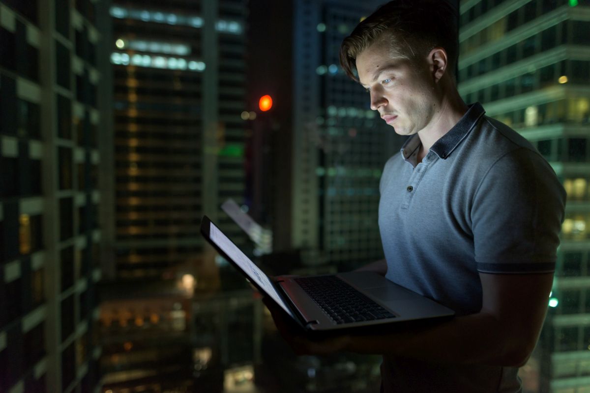 Man stands by a window at night using a laptop with city lights and tall buildings visible behind him. The screen casts a soft glow on his face, highlighting focused work in an urban setting.