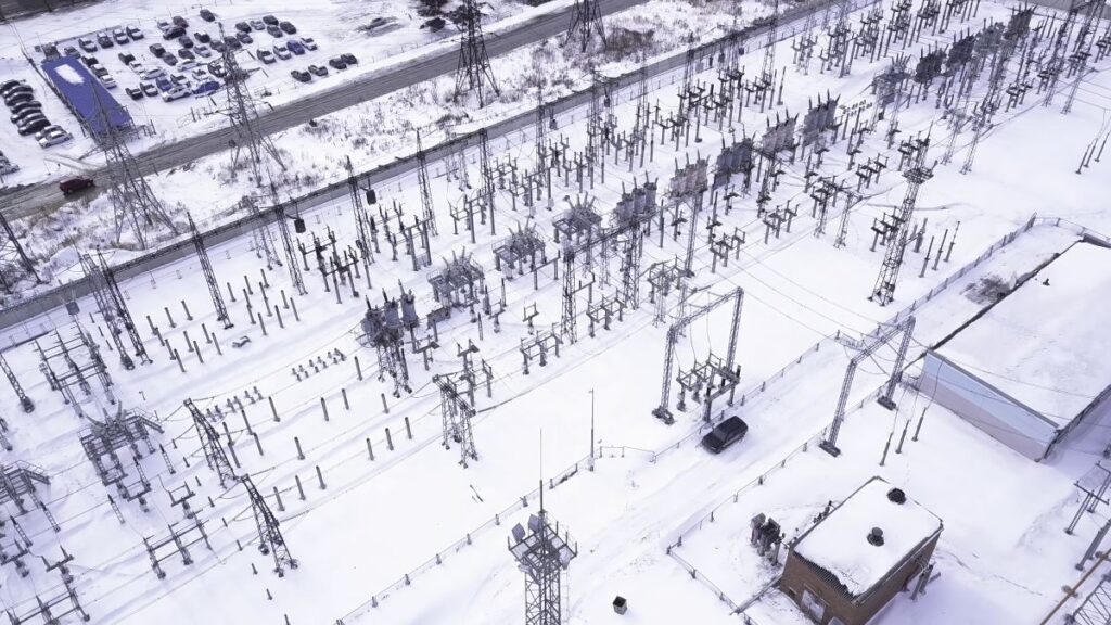 Aerial view of an electrical substation with transformers and power lines distributing high voltage, surrounded by snow-covered ground and buildings in a suburban area.