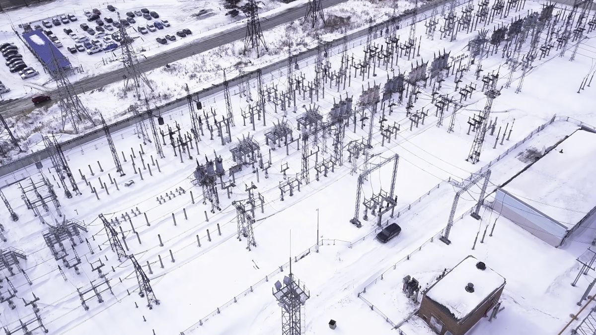 Aerial view of an electrical substation with transformers and power lines distributing high voltage, surrounded by snow-covered ground and buildings in a suburban area.