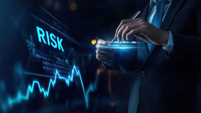 A suited business professional writes with a pen over a glowing digital interface labeled "RISK". A blue line graph and data visuals suggest regulatory risk analysis and risk management planning.
