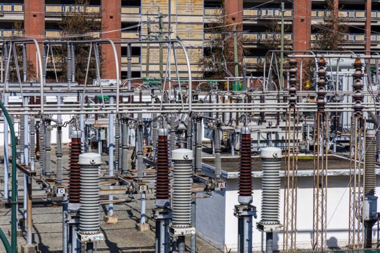 Urban electrical substation with insulators transformers and overhead power lines in front of brick apartment buildings.