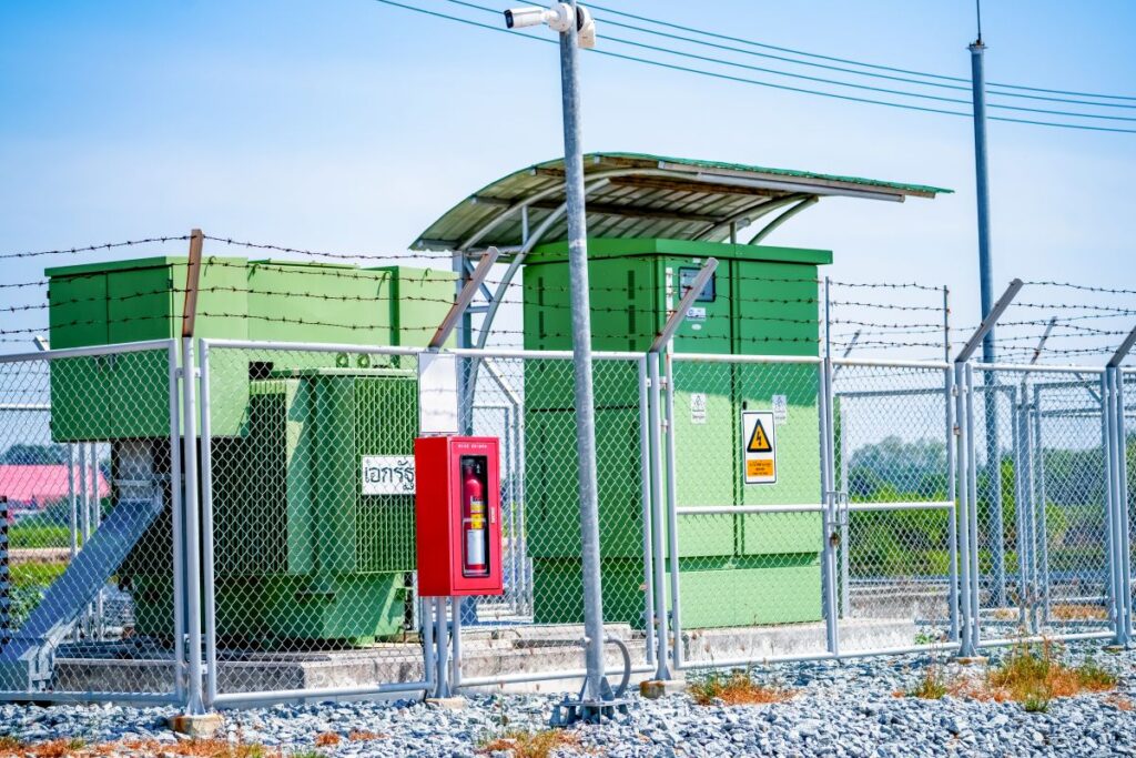 Green electrical power station equipment sits behind a chain link fence and barbed wire under a clear blue sky.