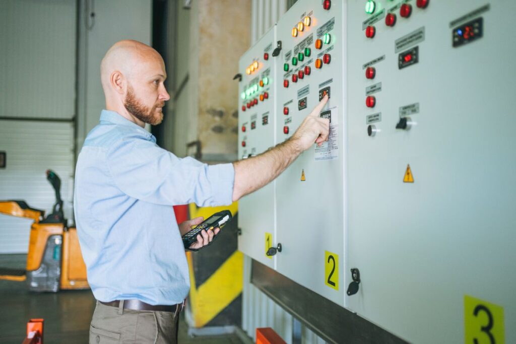 A technician presses an electrical control panel with red green and amber indicator lights while holding a handheld device.