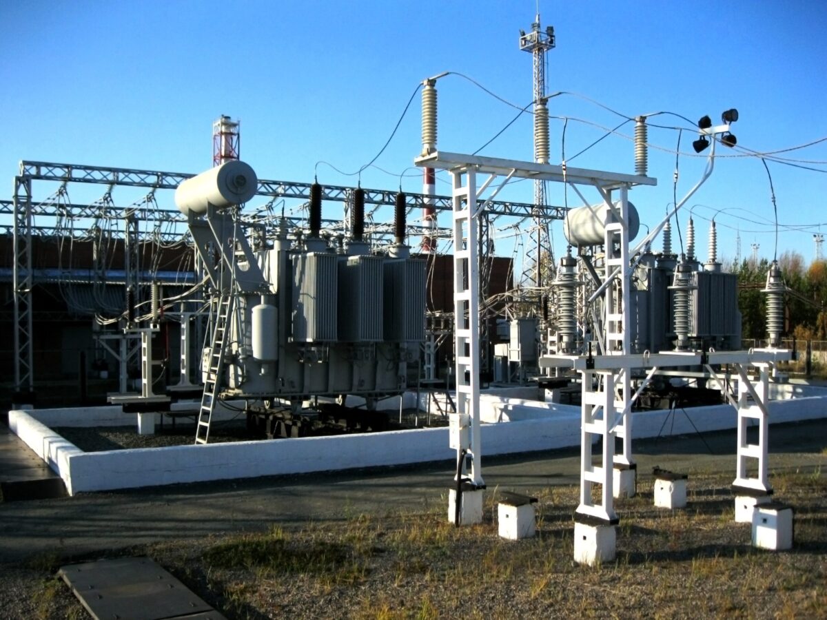 Electrical substation with transformers, insulators, and high-voltage equipment connected by overhead power lines under a clear blue sky. The industrial power grid infrastructure is surrounded by metal frameworks and transmission components used for electricity distribution and system reliability.