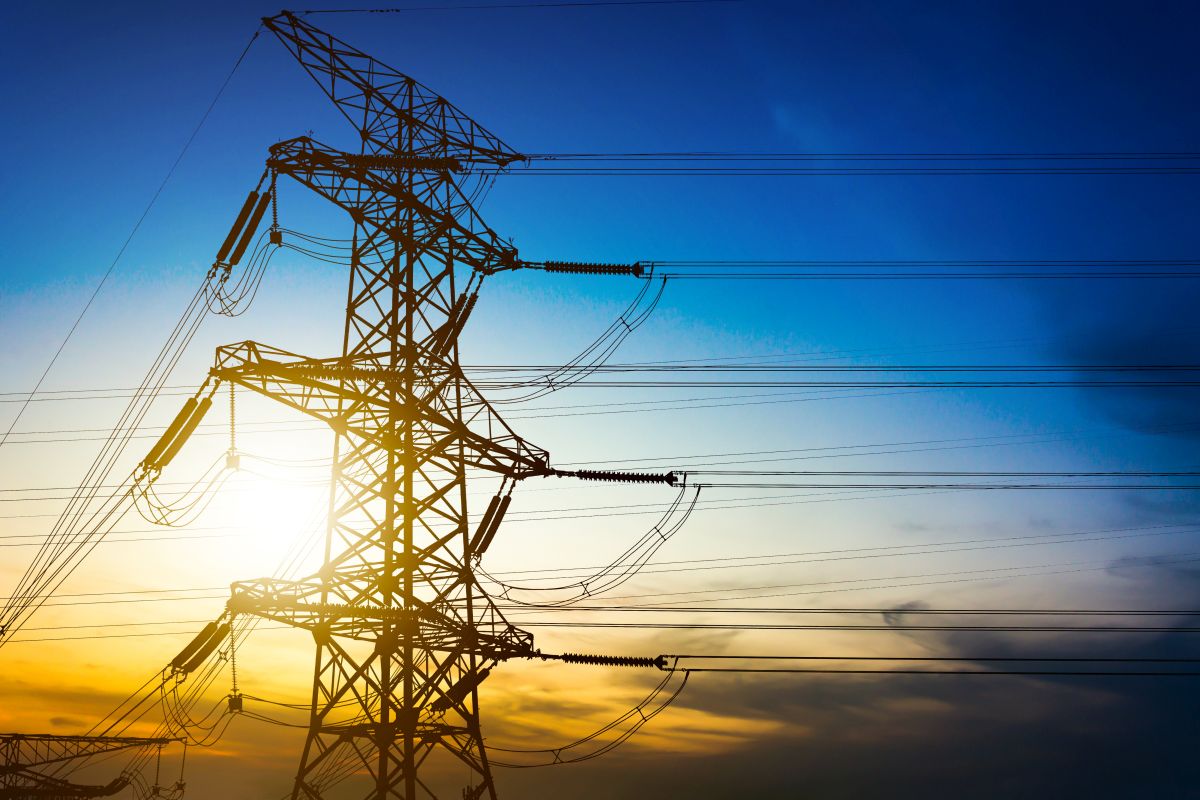 High voltage tower stands in silhouette against a blue and gold sunset sky with power lines extending across the frame.