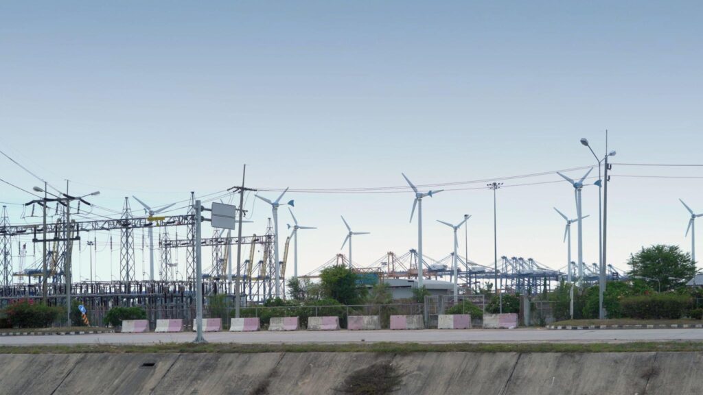 Electrical substation infrastructure with power lines and transformers in the foreground, alongside multiple wind turbines in the background near an industrial port, illustrating the integration of renewable energy with grid systems.