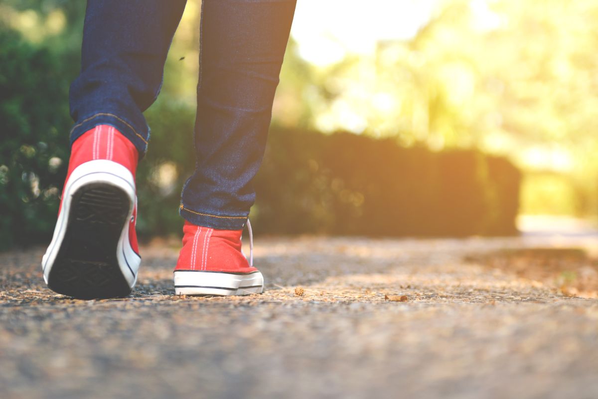 Close up view of a person walking along a park path wearing red sneakers and dark jeans, with sunlight glowing in the background.