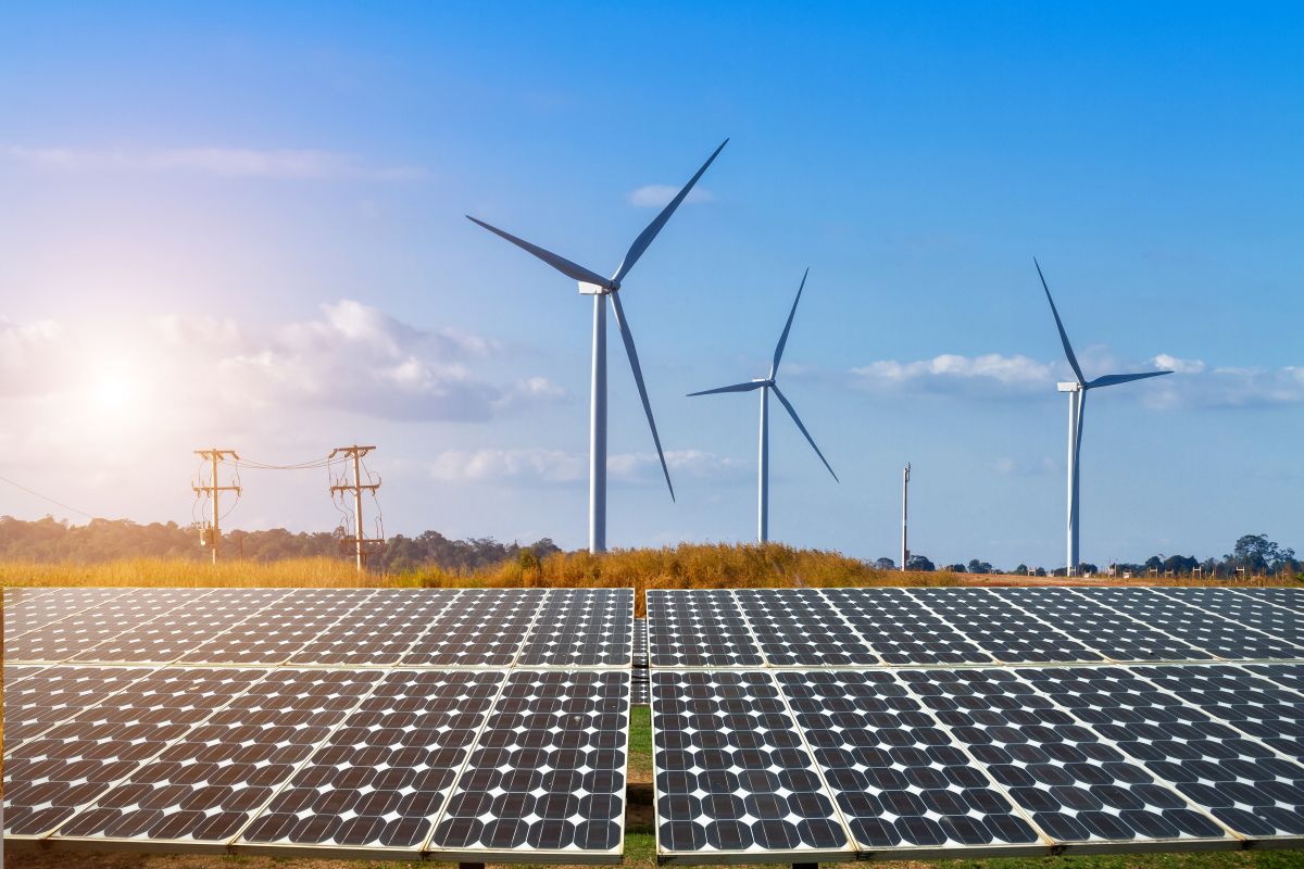 Solar panels in the foreground with wind turbines in the background, under a clear blue sky with sunlight.
