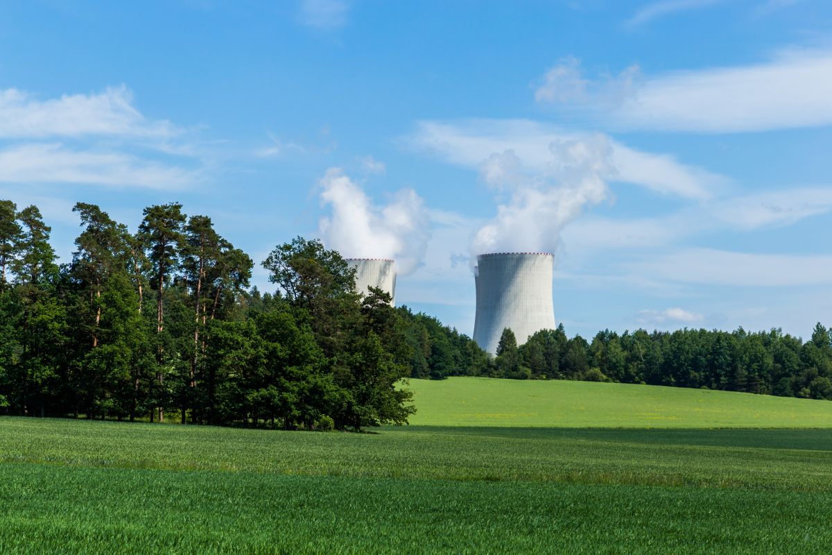 Nuclear plant cooling towers rising above a forest and green field under a blue sky.