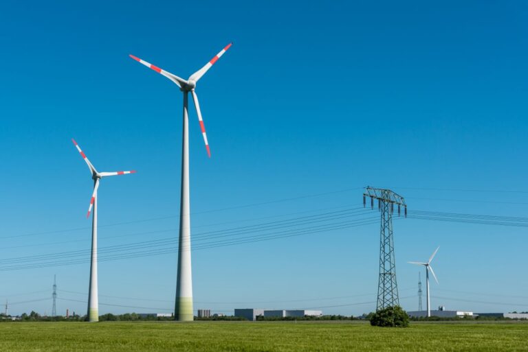 Two tall wind turbines with red tipped blades stand in a flat green field under a clear blue sky, with power lines and an electrical tower nearby showing wind energy generation in a rural landscape.