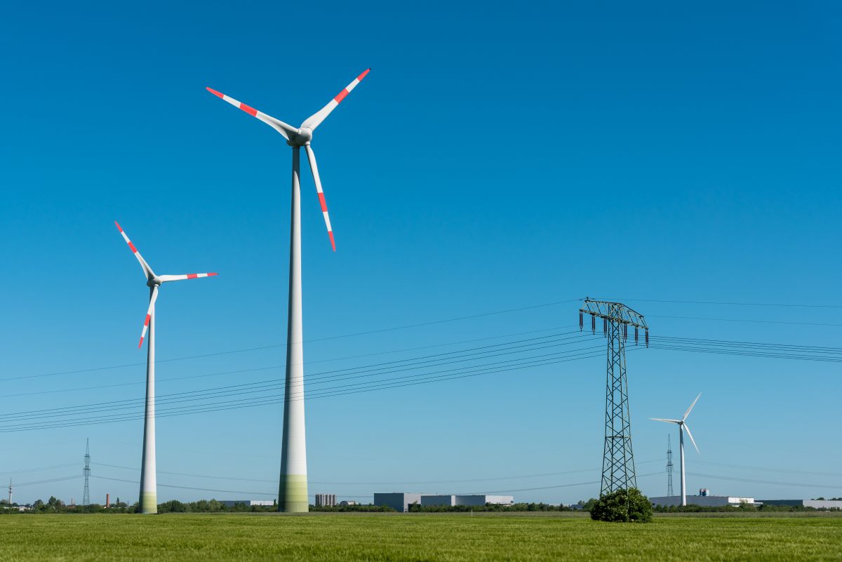 Two tall wind turbines with red tipped blades stand in a flat green field under a clear blue sky, with power lines and an electrical tower nearby showing wind energy generation in a rural landscape.