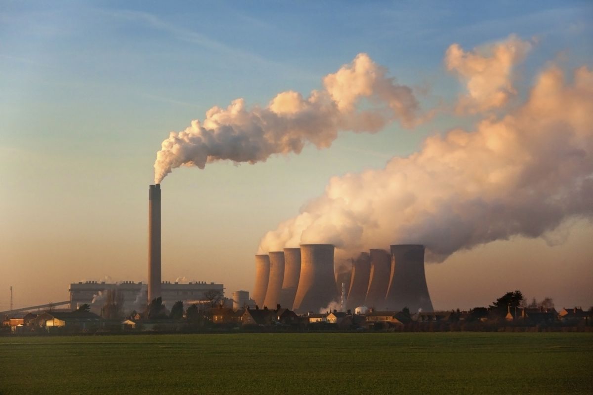A coal plant sits beyond a green field at sunset with a tall smokestack and cooling towers releasing thick clouds of steam into the sky.
