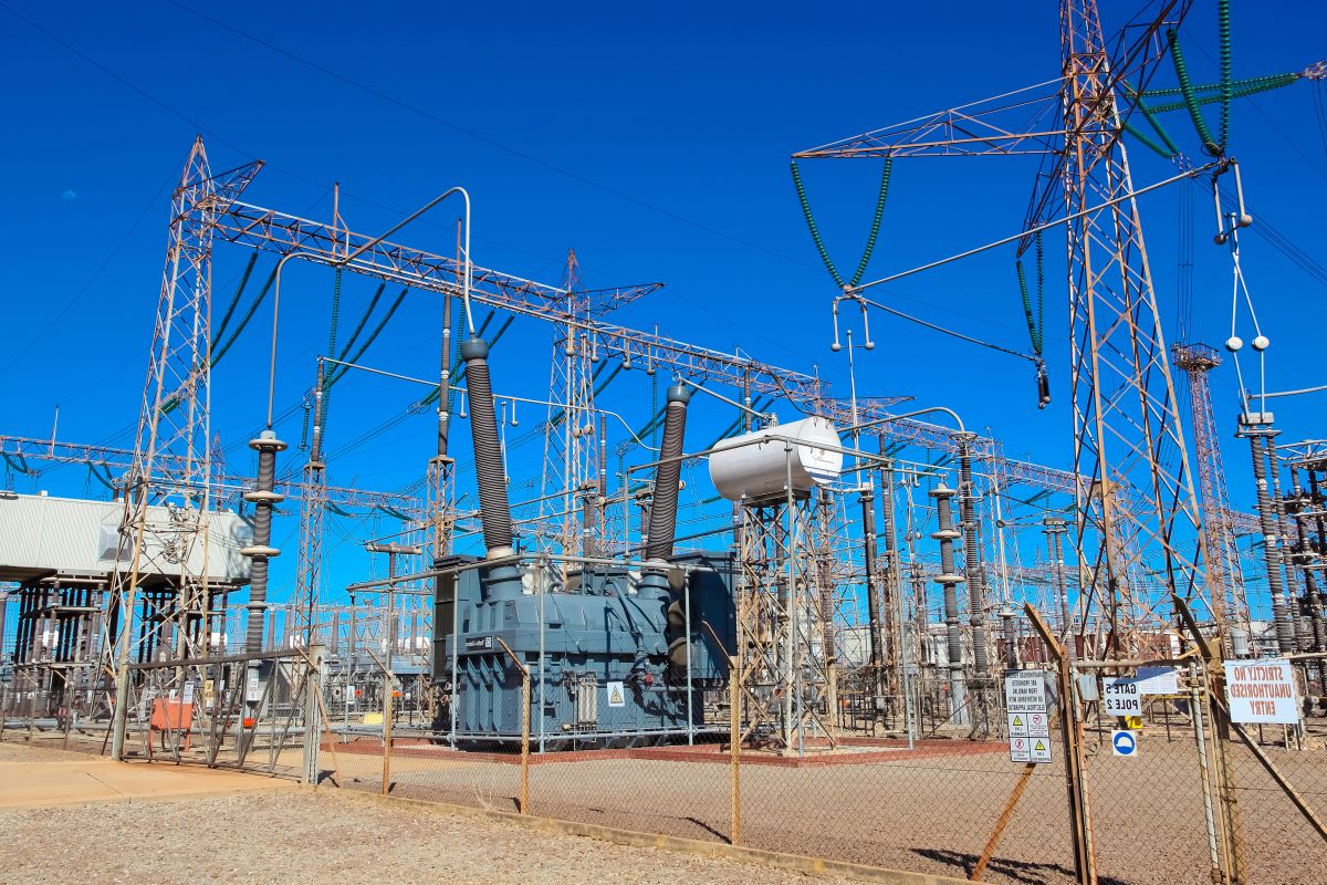 Electrical substation with transformers transmission towers and power lines under a bright blue sky.